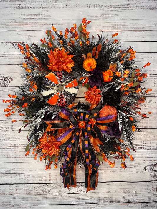 A Halloween-themed artificial wreath with orange flowers, small orange berries, dark green and silver faux foliage, a small pumpkin, and a large orange, black, and purple striped bow, displayed against a whitewashed wood plank background.