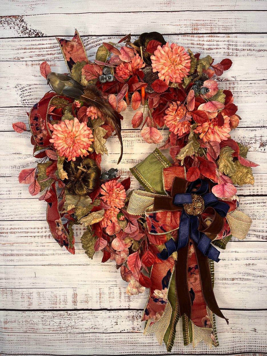 An autumn-themed fabric wreath with faux flowers, leaves, ribbons, and a small bird figure, displayed against a whitewashed wooden plank background.