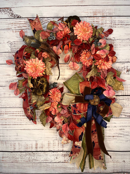 An autumn-themed fabric wreath with faux flowers, leaves, ribbons, and a small bird figure, displayed against a whitewashed wooden plank background.