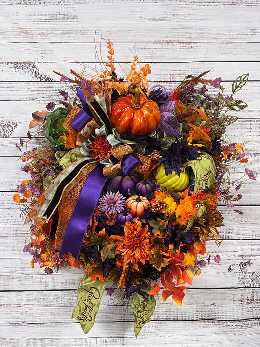 An autumn-themed wreath with faux pumpkins, orange and purple flowers, fall leaves, and a large purple and gold ribbon, displayed against a white-washed wood plank background.