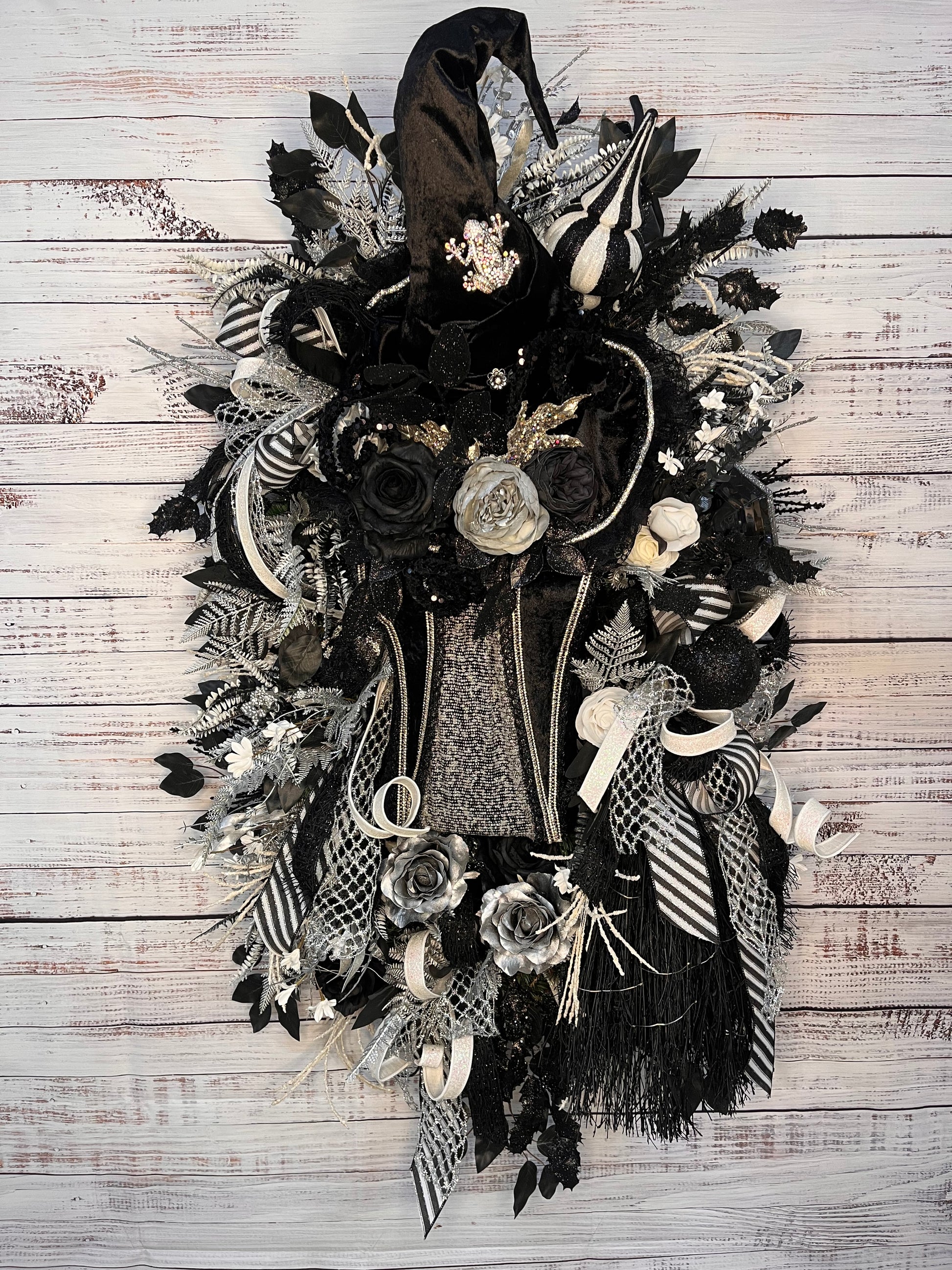 A gothic-themed decorative wreath featuring black fabric, ribbons, faux roses, tinsel, and silver-gray accents, displayed against a whitewashed wooden plank background.